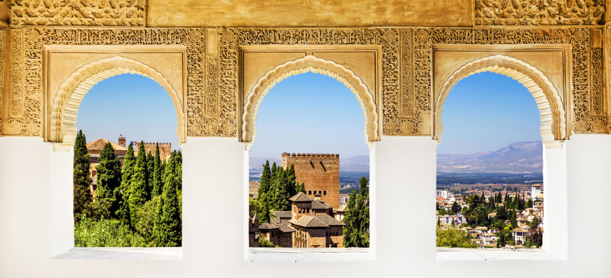 Ornate Moorish arches of the Alhambra in Granada frame a panoramic view of southern Spain's rolling hills, cypress trees, and historic towers. This image evokes the architectural splendor and rich Andalusian history featured in Vivida's Spain tour.