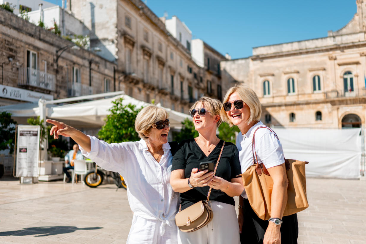 Three stylish women in their 50s and 60s share a joyful moment in a sun-drenched European square. This is the spirit of Vivida's tours: friendship, discovery, and the confidence of exploring beautiful destinations together